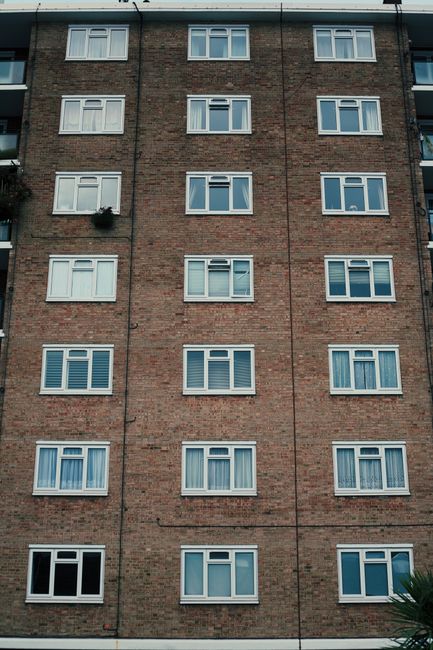 The image shows the exterior of a modern, multi-story residential building with a concrete facade featuring balconies with horizontal railings. The building has a somewhat weathered appearance with vi