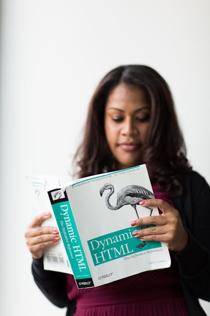 The image features a woman with shoulder-length dark hair, wearing a black blazer over a maroon top, standing indoors against a plain white wall. She is holding open a book titled 'Dynamic HTML' by O'
